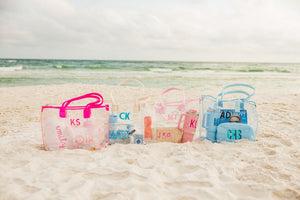 A group of clear totes are customized with monograms and are placed at the beach.