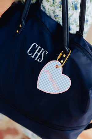 A navy blue tote bag with gold-tone hardware, featuring the monogram 'CHS' in white on the front and a heart-shaped light blue gingham key tag with pink script embroidery.