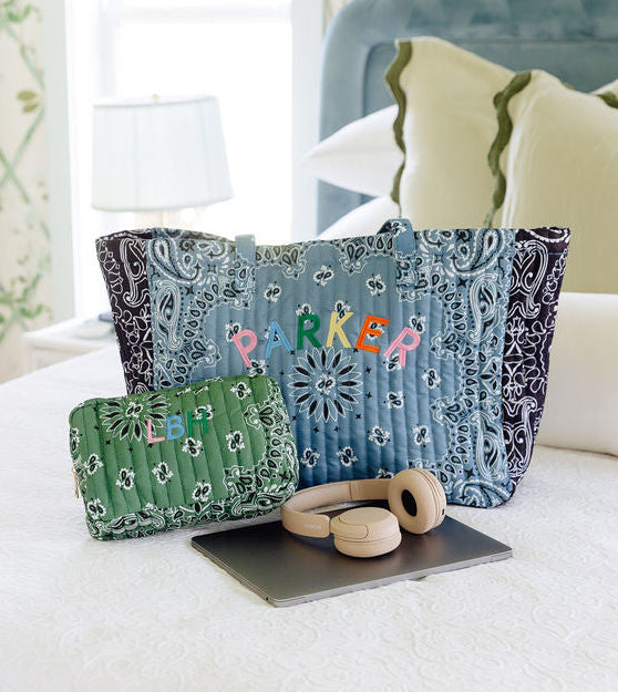 A blue and navy patterned tote bag with the name 'PARKER' embroidered in colorful letters sits on a white bed, alongside a matching green patterned pouch, wooden headphones, and a tablet. The background shows a softly lit bedroom with patterned curtains and pillows.