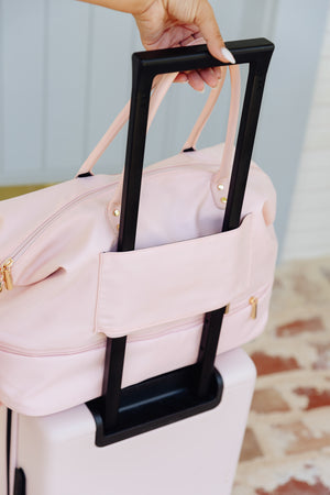 A light pink nylon duffel bag with a black handle and gold zipper pulls, shown with a white suitcase. A hand is holding the black handle of the bag, which is draped over the suitcase.