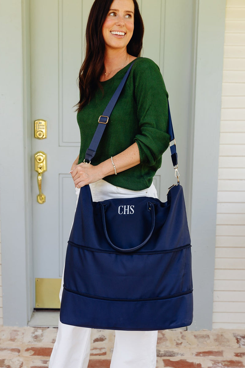 A woman stands on a front porch wearing a navy blue tote bag with white monogram lettering 'CHS' on the front pocket. The bag has a wide top opening, a top handle, and a shoulder strap.
