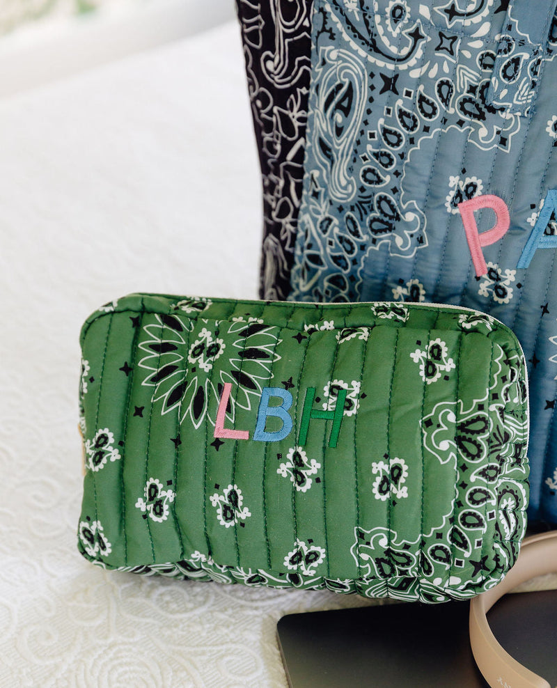 A green quilted bandana-patterned cosmetic pouch with colorful embroidered letters 'LBH' sits on a white textured surface next to a laptop, with other patterned bags in the background.