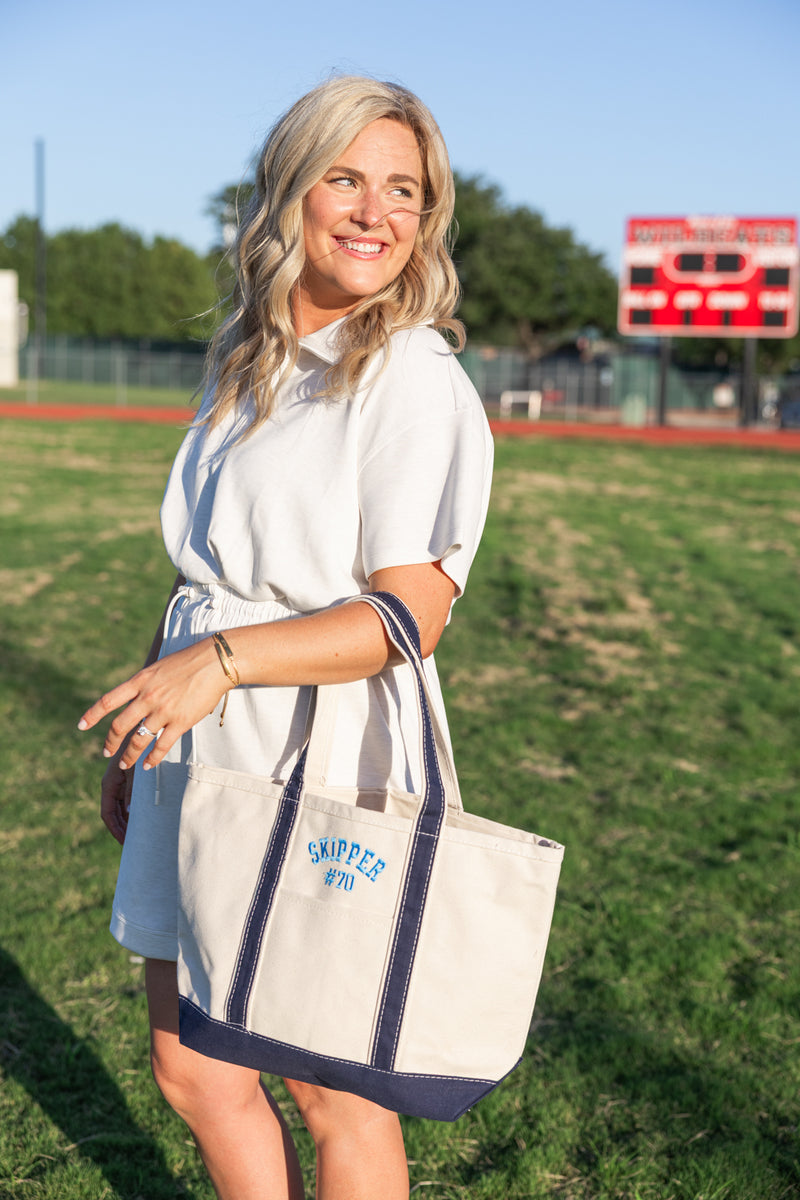 Woman holding natural canvas tote with navy trim and embroidered “SKIPPER #70” at a football field.