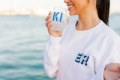 Smiling woman holding a personalized wine glass with blue monogram 'KL' while wearing a white sweatshirt embroidered with navy 'ER' initials, by the water.