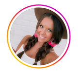 Woman in a hat with braided hair and bright pink earrings smiling outdoors against a white brick backdrop.
