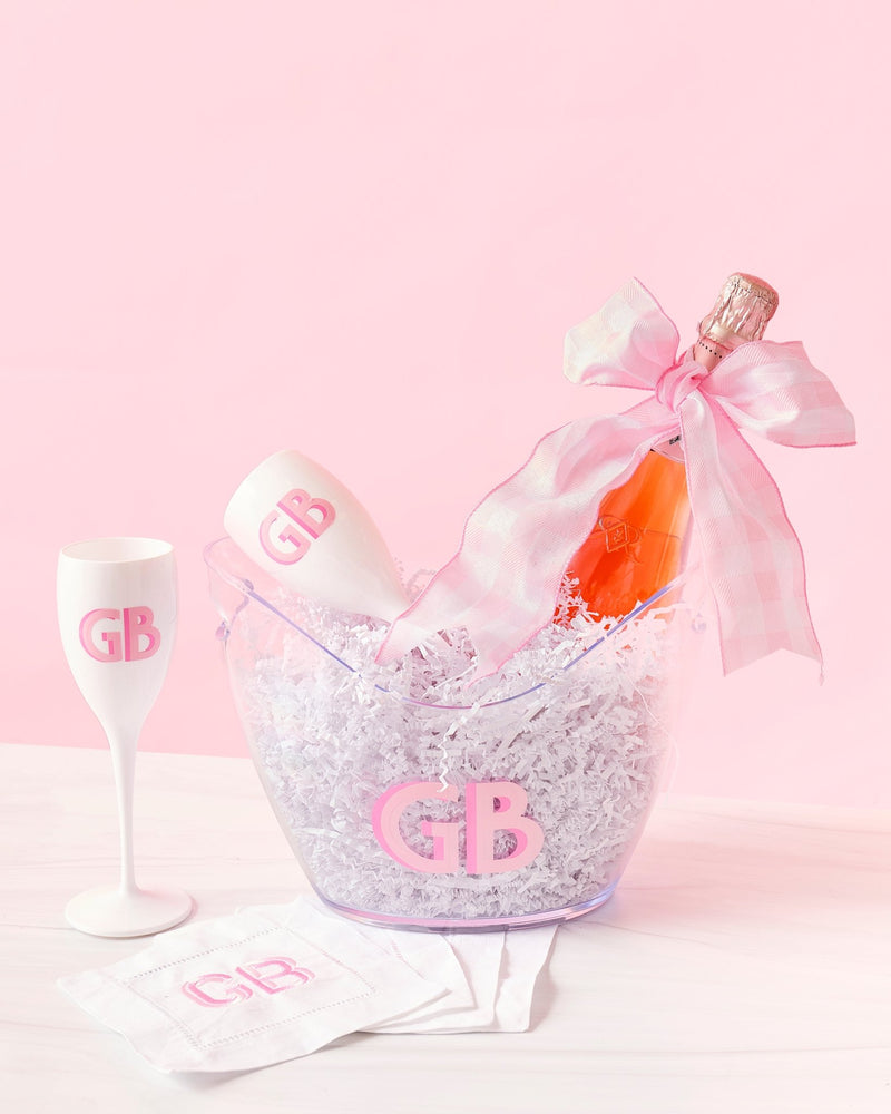 A set of white acrylic champagne flutes, a wide ice bucket and cocktail napkins are customized with a matching pink monogram.
