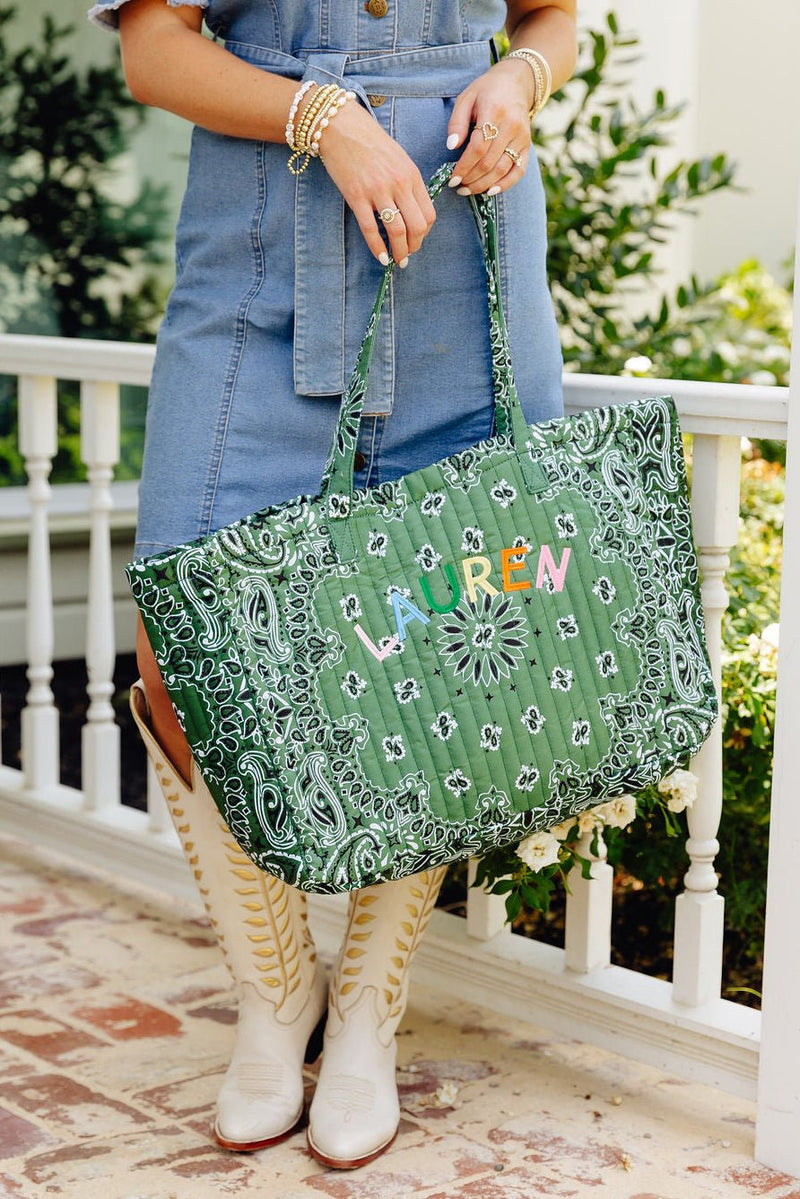 Woman holding a green bandana print quilted tote bag embroidered 'LAUREN' in rainbow letters, wearing white cowboy boots on a front porch