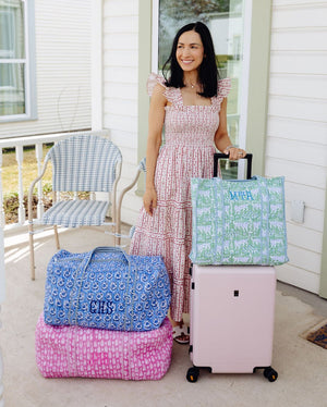 Woman standing with a pink suitcase, blue floral duffel bag monogrammed 'CHS', pink leaf print duffel bag, and green leopard print tote monogrammed 'MTA'