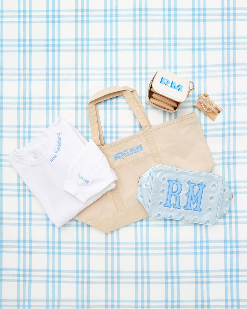 Personalized bride gift bundle featuring embroidered tote bag, custom monogram pouch, white t-shirt with blue embroidery, compact wallet, and keychain, displayed on a blue plaid background.