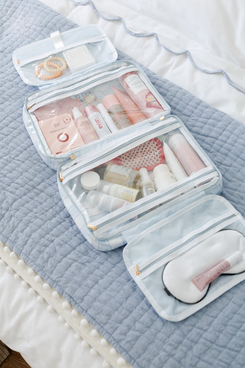 Open light blue striped clear coated hanging toiletry bag with skincare, hairbrush, and sleep mask organized in compartments on a bed