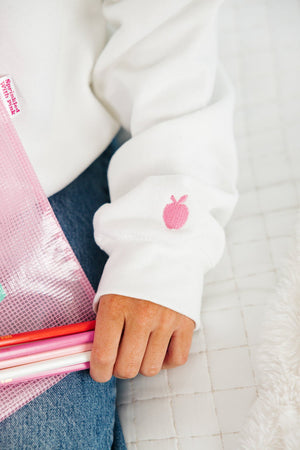 Close-up of a pink embroidered apple icon on the cuff of a white sweatshirt, with a person holding a clear pink zip pouch
