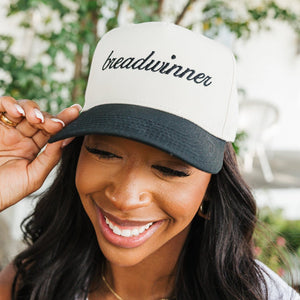 Image of a joyful woman adjusting a baseball cap with her right hand. The cap has a cream crown and a black bill, embroidered with the word 'breadwinner' in cursive black script. She is smiling broadly, showcasing her white tank top and delicate necklace, set against a blurred garden backdrop with lush greenery. Her expression reflects a playful and confident mood, emphasizing the empowering message on her cap.