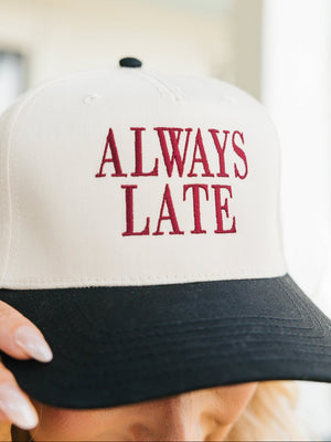 Close-up image of a baseball cap held by a person, visible only from the nose down. The cap has a white crown and a black bill, embroidered with the phrase 'ALWAYS LATE' in bold maroon letters.