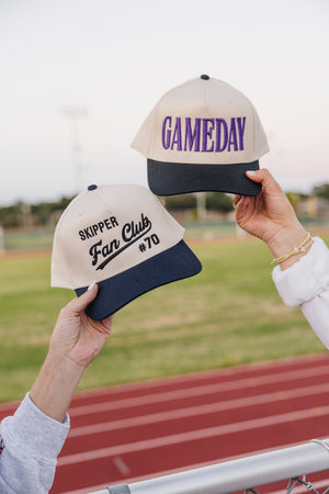 Hands lift featuring the purple-letter “GAMEDAY” cap beside another hat.