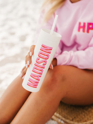 A girl on the beach holds a white skinny matte tumbler with her name in pink and orange font.