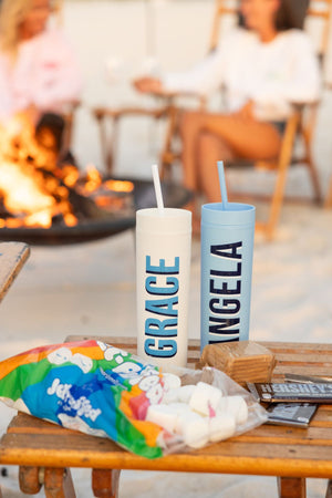 A blue and a white skinny matte tumbler are placed on a table with smores and customized with names.