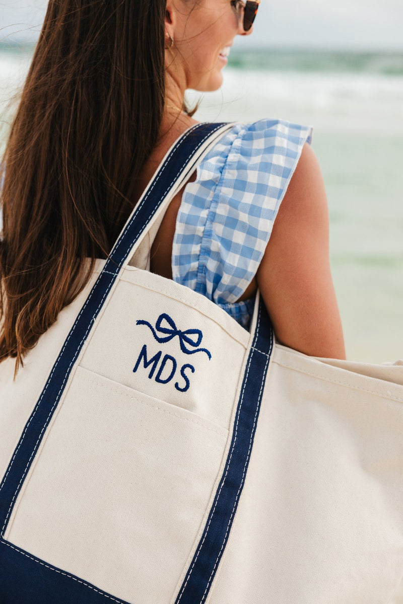 A girl at the beach in a blue dress holds a navy canvas tote with a navy embroidered bow and monogram on her shoulder
