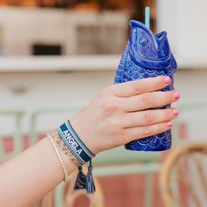 A person holds up a drink with a blue bracelet with a name embroidered on it in pink thread on their wrist