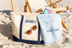 Personalized Embroidered Canvas Tote with navy blue straps and "DARNELL" stitched on the front, styled with sunglasses, a book, and a light blue embroidered towel reading "melissa"—perfect for a stylish beach day setup.