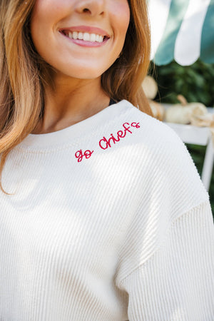 Woman wearing a cream corded sweatshirt embroidered with a personalized phrase on the collar and #87 on the sleeve, standing near a pumpkin display
