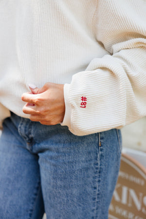 Close-up of a cream corded sweatshirt sleeve embroidered with #87 in red thread