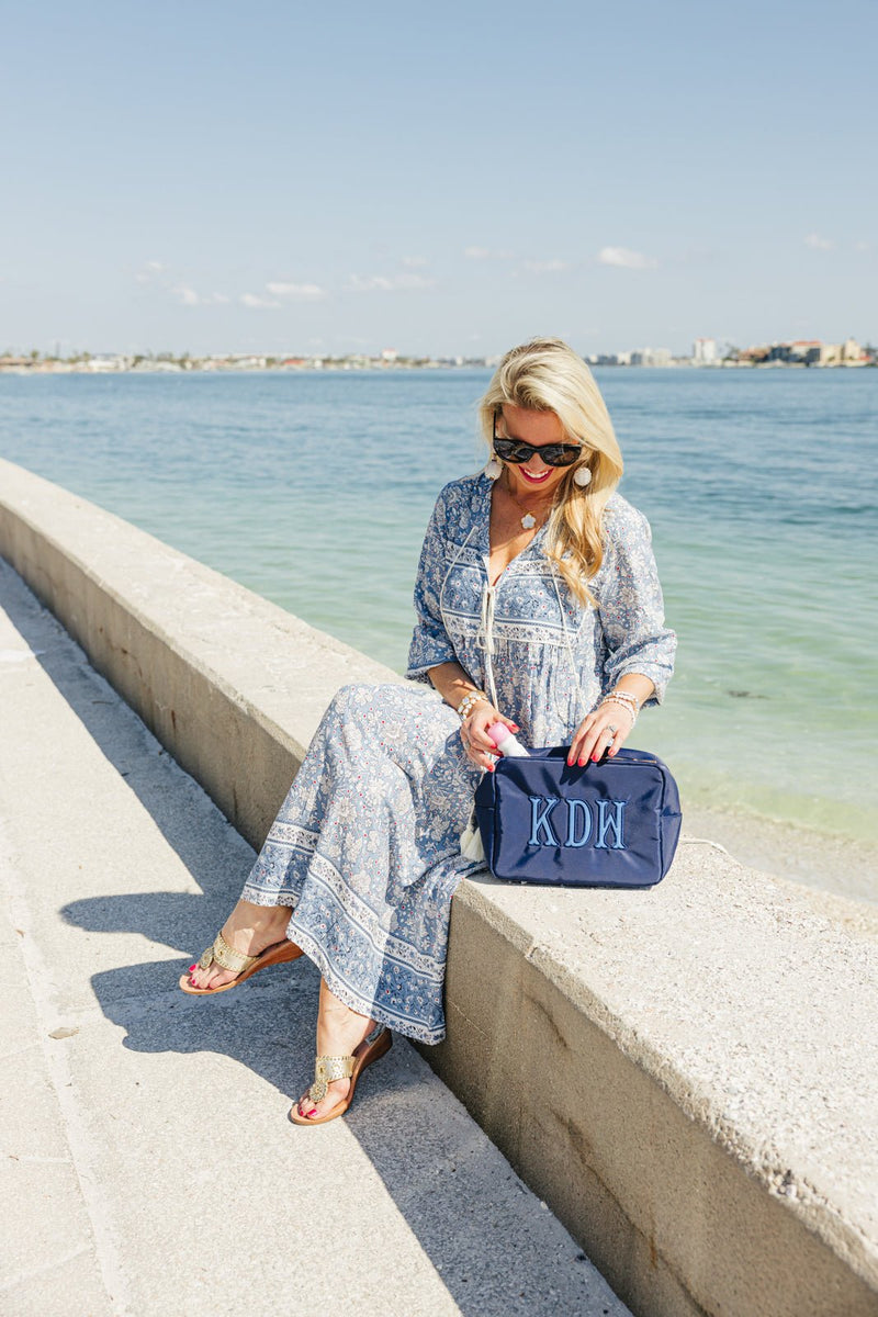 Woman in a blue floral dress sitting by the water holding a navy monogrammed travel bag with initials KDW.