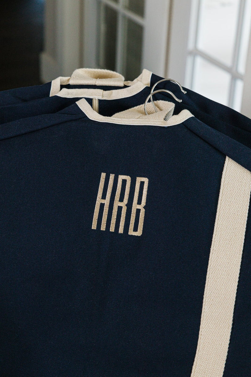 A navy blue personalized garment bag with embroidered initials "HRB" in beige thread hangs on a metal hanger, featuring a durable canvas material with a contrasting beige stripe detail. The elegant and functional travel accessory is designed to protect clothing while adding a stylish, monogrammed touch. The bag is part of a matching set, ideal for coordinated travel organization.