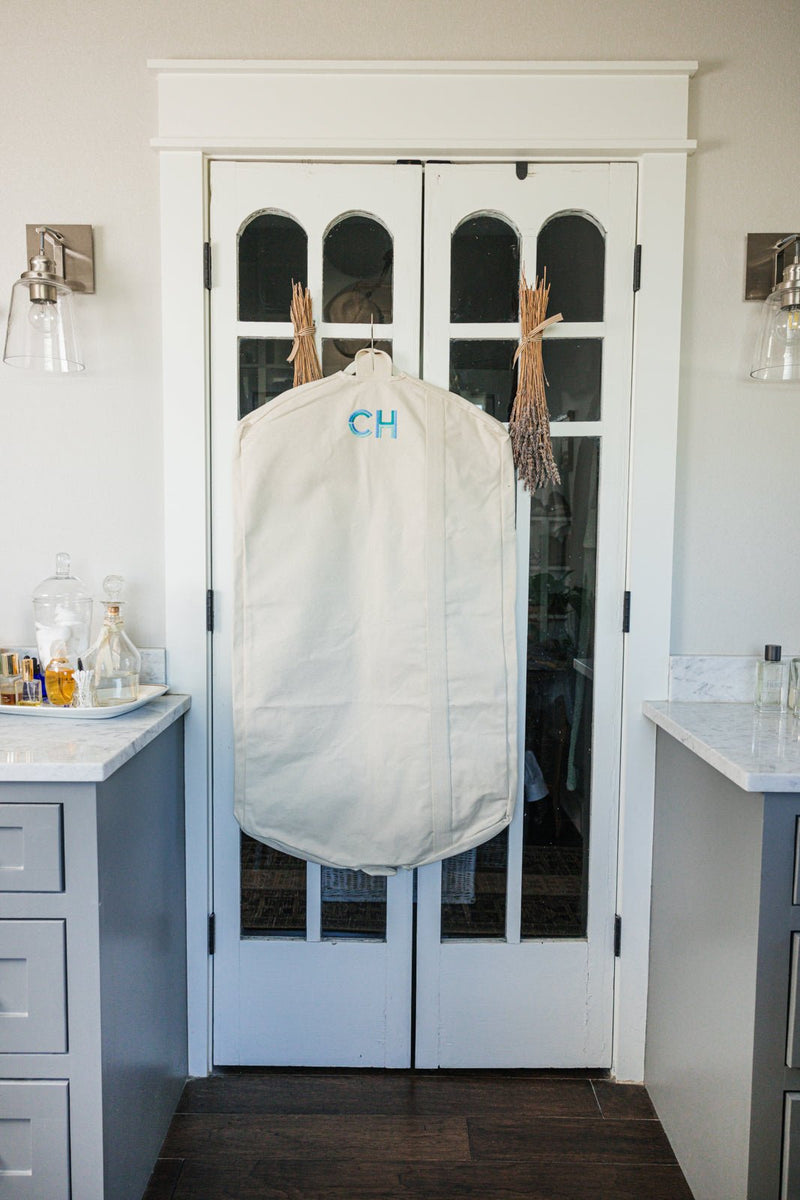 A personalized beige garment bag with embroidered initials "CH" hangs elegantly on a set of French doors in a modern home. The high-quality canvas bag is designed for stylish and organized travel, protecting clothing while adding a touch of customization. The setting features a neutral-toned interior with sophisticated decor, including sconces, marble countertops, and dried floral accents, creating a refined and inviting atmosphere.