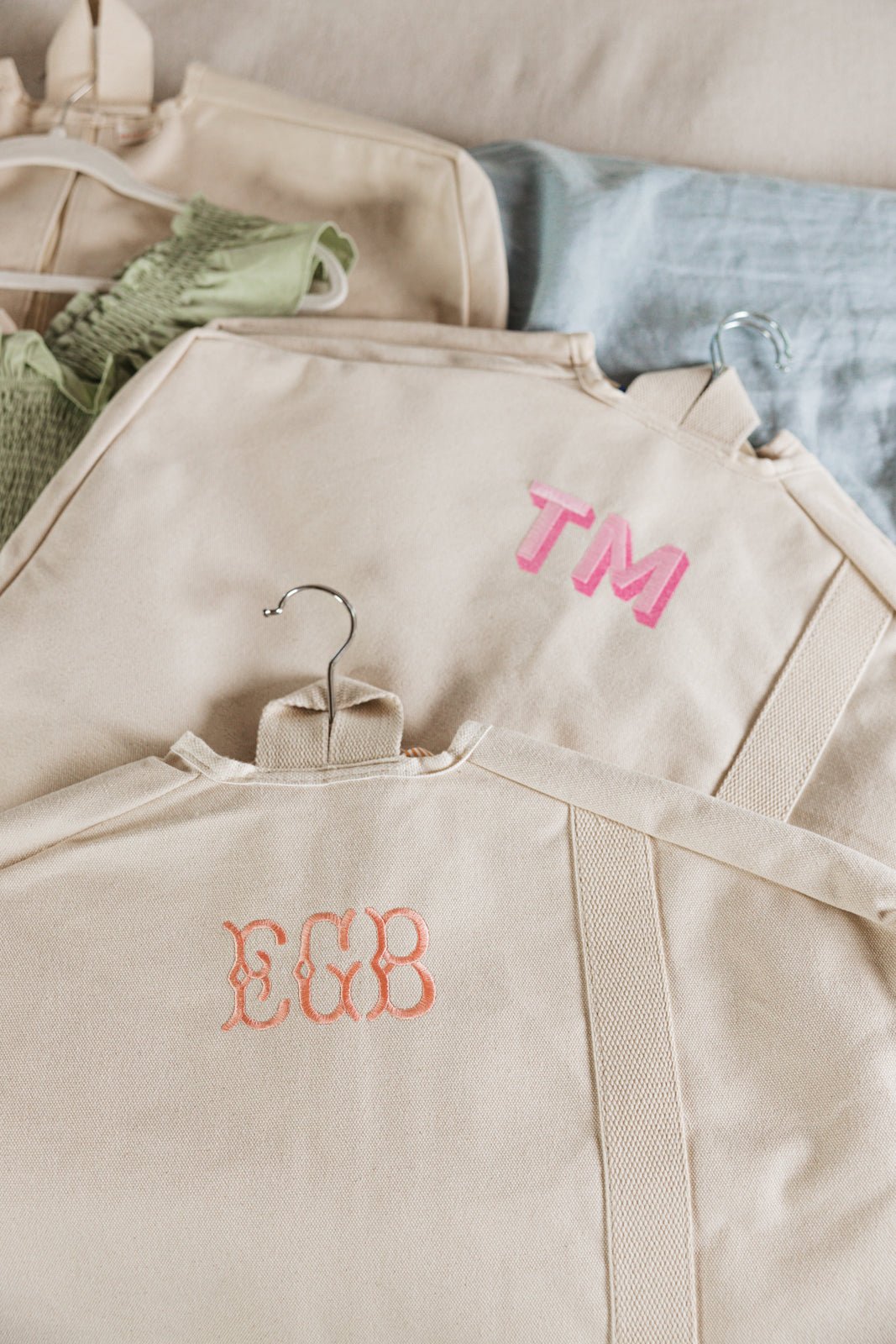 A stylish set of two personalized garment bags in navy and beige, each featuring custom monogram embroidery. The navy bag showcases "NDW" in light blue thread, while the beige bag is adorned with "SN" in soft pink. Designed for travel and organization, these canvas garment bags offer a chic and functional way to protect clothing while on the go.