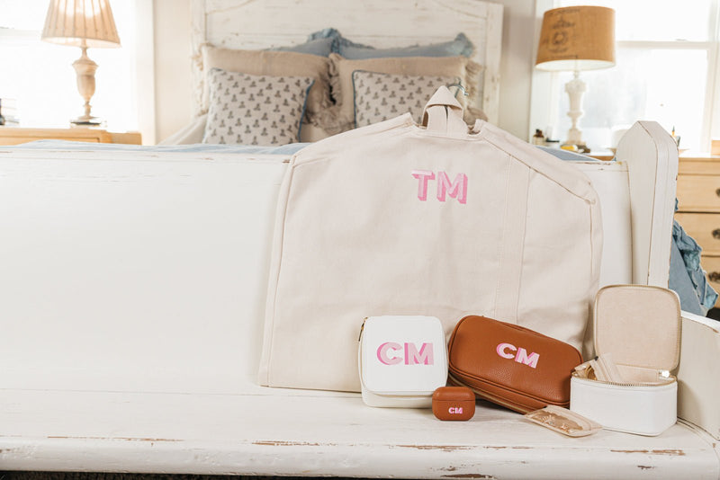 A beautifully coordinated personalized travel set is displayed on a white wooden bench in a cozy bedroom setting. The set includes a monogrammed beige canvas garment bag with "TM" embroidered in pink, a white travel jewelry case with "CM" in pink, a brown leather tech organizer with matching "CM" embroidery, a compact white jewelry box, and a brown monogrammed AirPods case. A small jewelry pouch with gold accessories is also included, enhancing the set's elegance.