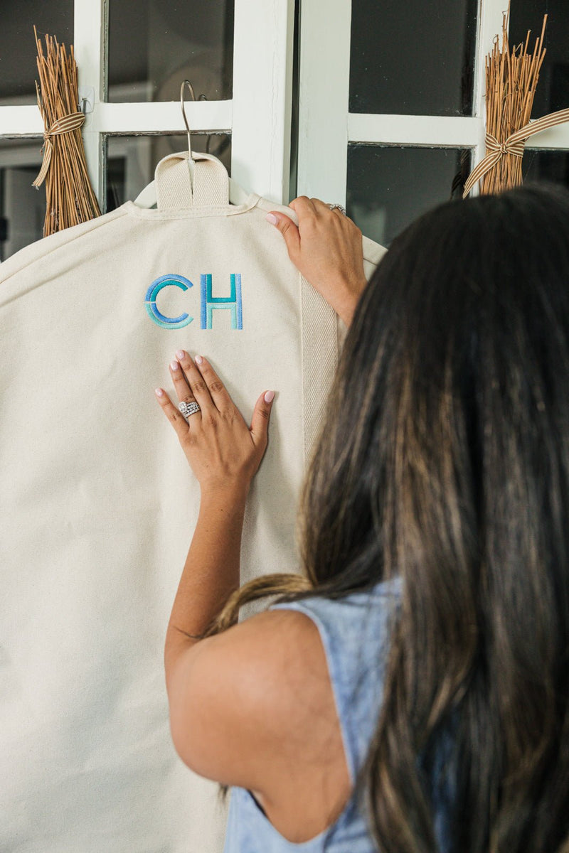 A woman with long dark hair and a denim dress gently places her hands on a beige personalized garment bag with embroidered initials "CH" in a blue gradient thread. The canvas garment bag, part of a coordinated travel set, hangs neatly on a door with decorative wheat accents, showcasing its durable design and stylish monogram.