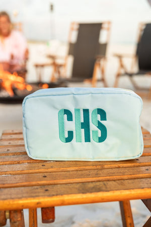 A blue nylon pouch is customized with a mint and turquoise monogram and placed on a table by a campfire.
