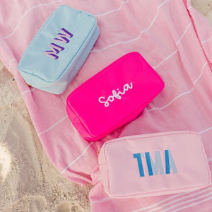 A group of large nylon pouches ate customized with embroidered names and monograms and are laid out on a pink turkish towel.