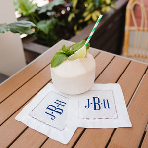 A set of cocktail napkins are embroidered with a navy monogram and placed under a coconut drink.