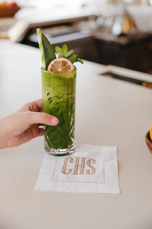 A cocktail napkin is an embroidered with a beige monogram and placed under a green drink.