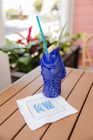 A cocktail napkin is an embroidered with a blue monogram and placed under a drink in a blue fish cup.