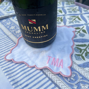 A chilled bottle of Mumm sparkling wine rests on a light blue Embroidered Monogram Scalloped Cocktail Napkin with pink trim and personalized “TMA” initials, part of a set of 4 elegant monogrammed napkins, displayed on a patterned outdoor tablecloth.