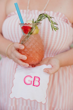 A woman in a pink striped dress holds a fruity cocktail and a pink scalloped cocktail napkin with a pink monogram.