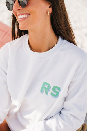 A girl smiles in a white sweatshirt with a mint and blue embroidered monogram.