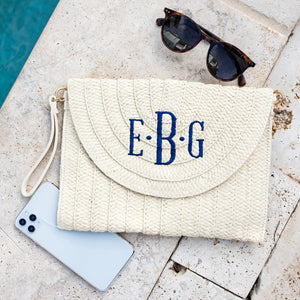 A straw clutch with a navy embroidered monogram is placed by a pool next to a pair of sunglasses and a phone