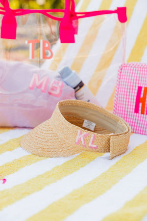 A straw visor is placed on a yellow cabana towel and is customized with a pink monogram.