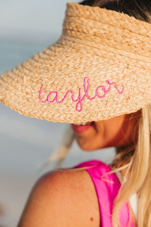 A woman wears a straw visor with an embroidered name in pink thread.