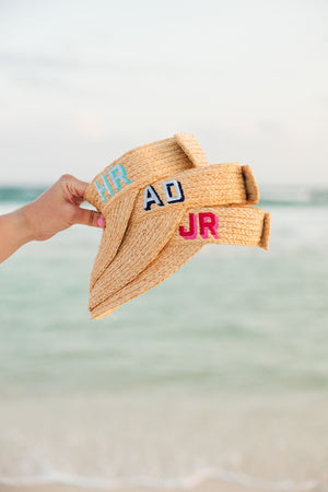A person holds up three straw visors with colorful monograms.