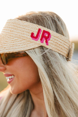 A woman wears a straw visor with a red and pink embroidered monogram.
