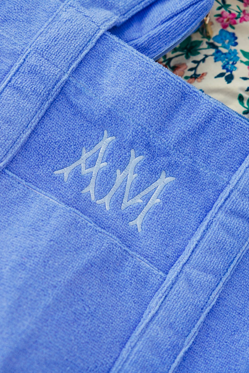 Close-up of a personalized blue terry cloth tote bag from Sprinkled with Pink, featuring an elegant monogrammed "AM" embroidery in a stylish script font. The custom beach tote is made of soft, absorbent fabric, making it perfect for carrying essentials to the pool, beach, or on summer travels. The floral fabric in the background enhances the chic and relaxed vacation aesthetic, showcasing this monogrammed tote set as both fashionable and functional.