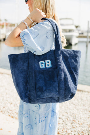 A stylish woman in a light blue eyelet dress carries a navy blue personalized terry cloth tote bag embroidered with the initials "GB" in bold, varsity-style lettering. The plush texture and oversized design make this custom beach tote from Sprinkled with Pink perfect for boating, beach trips, and summer getaways. Captured by the waterfront with yachts in the background, this monogrammed tote blends practicality and coastal elegance, offering a chic and functional accessory for travel or everyday use.