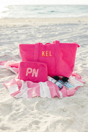 A vibrant pink personalized tote bag and matching cosmetic pouch set from Sprinkled with Pink, elegantly displayed on a sandy beach. The tote bag, embroidered with "KEL", features sturdy handles and ample storage for beach day essentials, while the cosmetic pouch, monogrammed with "PN", is perfect for organizing smaller items. Set on a stylish pink and white striped beach towel alongside sunglasses, sunscreen, and a magazine, this set is ideal for vacations, bridesmaid gifts, or a chic everyday carryall.
