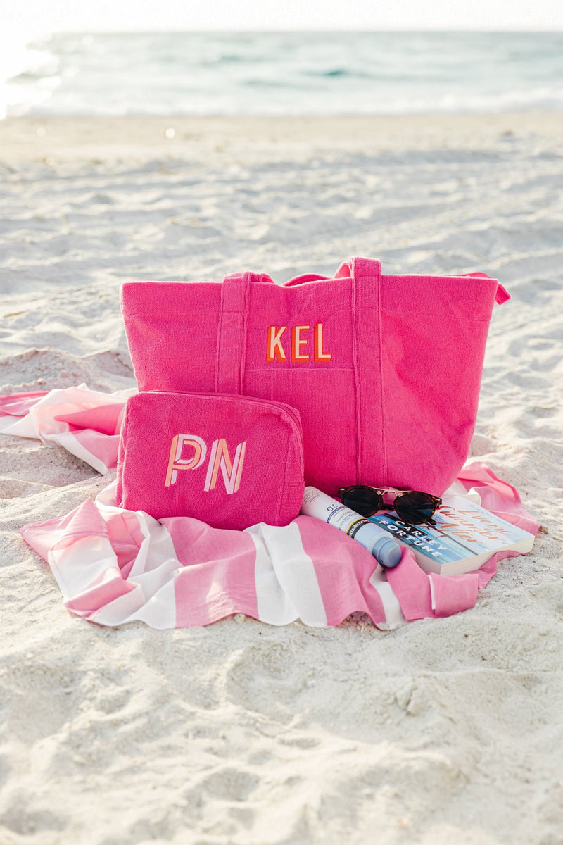 A vibrant pink personalized tote bag and matching cosmetic pouch set from Sprinkled with Pink, elegantly displayed on a sandy beach. The tote bag, embroidered with "KEL", features sturdy handles and ample storage for beach day essentials, while the cosmetic pouch, monogrammed with "PN", is perfect for organizing smaller items. Set on a stylish pink and white striped beach towel alongside sunglasses, sunscreen, and a magazine, this set is ideal for vacations, bridesmaid gifts, or a chic everyday carryall.