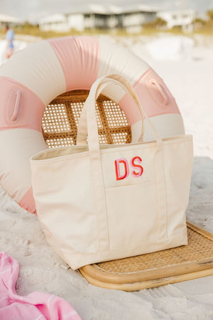 A cream coated canvas tote is customized with a pink and orange monogram and placed on the beach with a pink floatie.