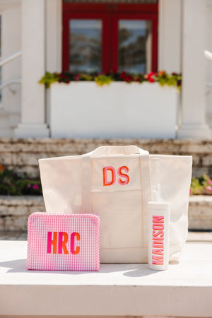 A clear coated canvas tote is customized with an orange and pink monogram and placed with a pink roadie and white skinny tumbler.